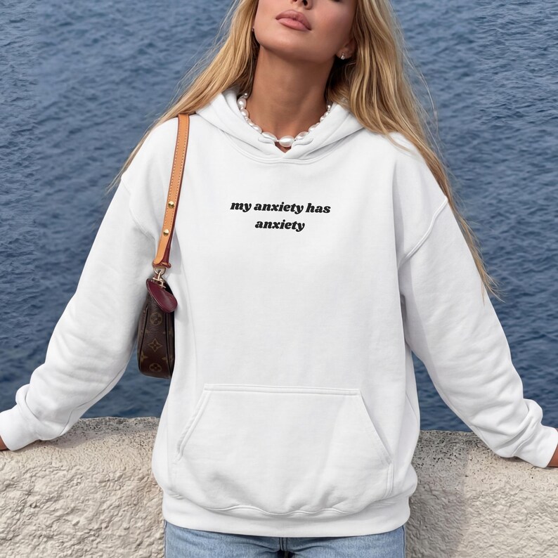 Peut inclure: Un sweat-shirt &agrave; capuche blanc &agrave; manches longues avec le texte noir "my anxiety has anxiety" imprim&eacute; sur la poitrine. Le v&ecirc;tement comprend une poche kangourou &agrave; l'avant, une capuche &agrave; cordon et des poignets c&ocirc;tel&eacute;s, avec un sac &agrave; bandouli&egrave;re marron.
