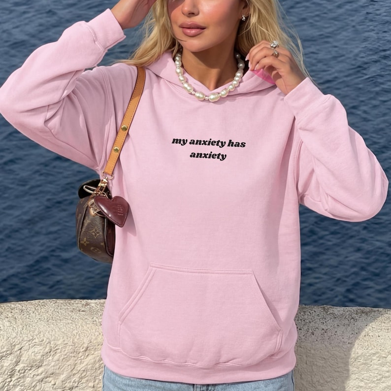Peut inclure: Un sweat-shirt &agrave; capuche rose clair &agrave; manches longues avec une poche kangourou sur le devant et le texte noir "my anxiety has anxiety" imprim&eacute; sur la poitrine. Le v&ecirc;tement est port&eacute; avec un sac &agrave; bandouli&egrave;re marron et un collier de perles.