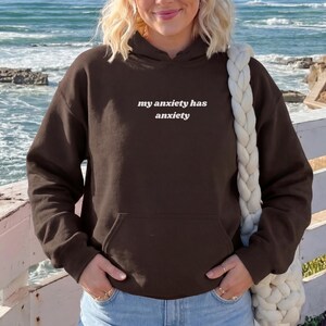 Peut inclure: Un sweat-shirt &agrave; capuche marron fonc&eacute; &agrave; manches longues avec le texte blanc "my anxiety has anxiety" imprim&eacute; sur la poitrine. Le v&ecirc;tement comprend une poche kangourou sur le devant, des manches longues &agrave; poignets c&ocirc;tel&eacute;s et une capuche assortie.