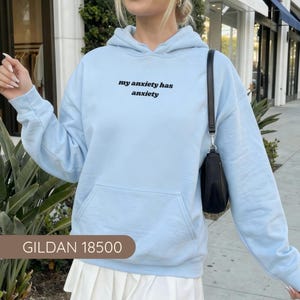 Peut inclure: Un sweat-shirt &agrave; capuche bleu clair avec le texte noir "my anxiety has anxiety" imprim&eacute; sur la poitrine. Le v&ecirc;tement comprend une poche kangourou &agrave; l'avant, des manches longues avec poignets c&ocirc;tel&eacute;s et une capuche &agrave; cordon. Un sac noir est port&eacute;.