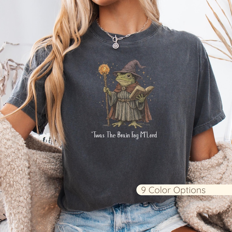 A person is wearing a vintage-wash charcoal gray T-shirt featuring a whimsical fantasy-style illustration of a frog dressed as a wizard. The frog wears a pointed hat and robe, holds a glowing staff in one hand, and an open book in the other. Beneath the graphic, the text reads “’Twas The Brain Fog M’Lord.” The outfit is styled with blue jeans, a silver necklace, and a cozy beige sherpa jacket draped off the shoulders. A “9 Color Options” label appears in the lower-right corner.