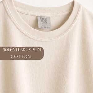K&ouml;nnte beinhalten: Cremefarbenes T-Shirt mit Rundhalsausschnitt. Das Etikett lautet "Comfort Colors" und "XL - TG - EG". Der Text "100% RING SPUN COTTON" ist auf einem braunen rechteckigen Etikett zu sehen. Das Shirt besteht aus weicher Baumwolle.