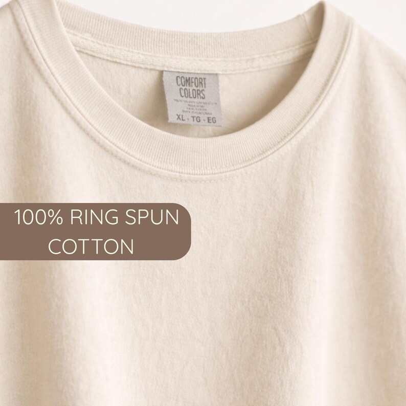 K&ouml;nnte beinhalten: Cremefarbenes T-Shirt mit Rundhalsausschnitt. Das Etikett lautet "Comfort Colors" und "XL - TG - EG". Der Text "100% RING SPUN COTTON" ist auf einem braunen rechteckigen Etikett zu sehen. Das Shirt besteht aus weicher Baumwolle.