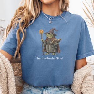 K&ouml;nnte beinhalten: Blaues T-Shirt mit einer Illustration eines Froschzauberers, der einen Stab und ein Buch h&auml;lt und einen Hut und einen Umhang tr&auml;gt. Der Text "'Twas The Brain Fog M'Lord" ist darunter gedruckt. Das T-Shirt hat kurze &Auml;rmel.
