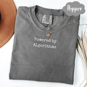May include: A gray t-shirt with the text "Powered by Algorithms" in white. The shirt has a crew neck and short sleeves. A silver necklace with a round pendant is draped across the shirt.