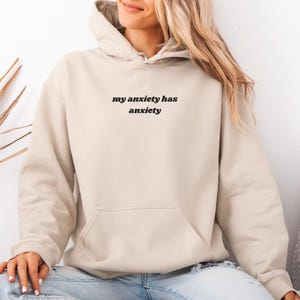 Peut inclure: Un sweat-shirt &agrave; capuche beige avec le texte noir "my anxiety has anxiety" imprim&eacute; au centre de la poitrine. Le v&ecirc;tement comprend une capuche avec des cordons de serrage et une grande poche kangourou &agrave; l'avant, fait d'un tissu uni.