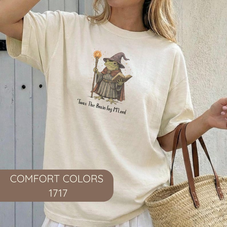 K&ouml;nnte beinhalten: Cremefarbenes T-Shirt mit einer skurrilen Illustration eines Froschzauberers, der einen Stab und ein Buch h&auml;lt, mit dem Text "'Twas The Brain Fog M'Lord". Das Shirt hat auch den Text "COMFORT COLORS 1717" unten.