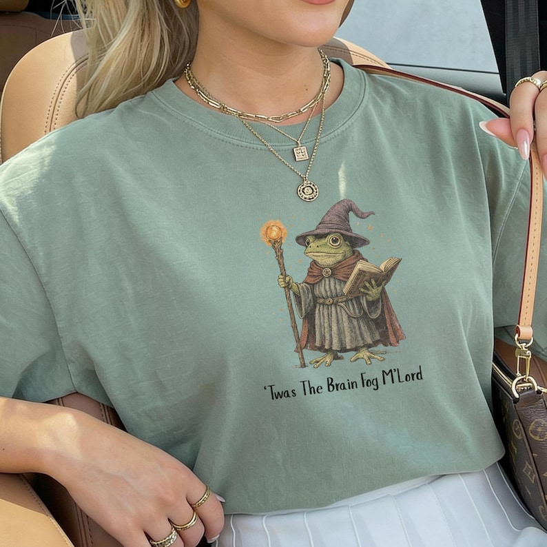 K&ouml;nnte beinhalten: Salbeigr&uuml;nes T-Shirt mit einer Illustration eines Froschzauberers, der einen leuchtenden Stab und ein Buch h&auml;lt. Der Frosch tr&auml;gt einen Zaubererhut und einen Umhang. Der Text "Twas The Brain Fog M'Lord" ist darunter gedruckt.