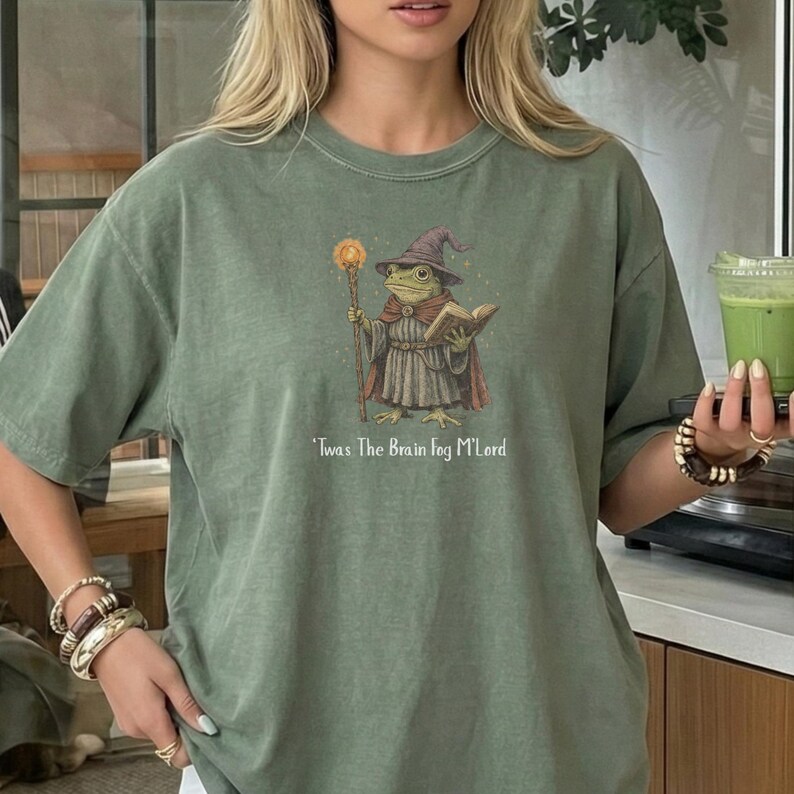 K&ouml;nnte beinhalten: Salbeigr&uuml;nes T-Shirt mit einer Illustration eines Froschzauberers. Der Frosch tr&auml;gt einen lila Zaubererhut und Umhang und h&auml;lt einen Stab und ein Buch. Der Text "'Twas The Brain Fog M'Lord" ist darunter gedruckt. Ein skurriles Design.