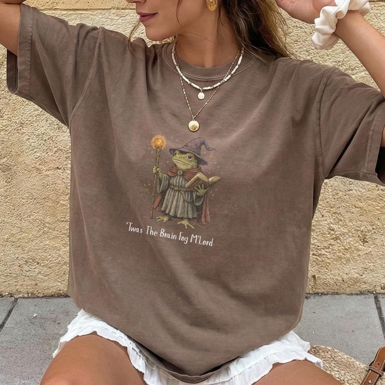 K&ouml;nnte beinhalten: Braunes T-Shirt mit einer Illustration eines Froschzauberers, der einen Stab und ein Buch h&auml;lt, mit dem Text "Twas The Brain Fog M'Lord". Der Frosch tr&auml;gt einen Zaubererhut und -robe. Kurz&auml;rmlig.