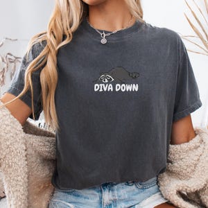 Puede incluir: Una camiseta de manga corta en color gris carbón que presenta un gráfico de un mapache tumbado con el texto DIVA DOWN impreso en letras blancas debajo. La prenda tiene cuello redondo y un corte holgado con un acabado de aspecto desgastado.