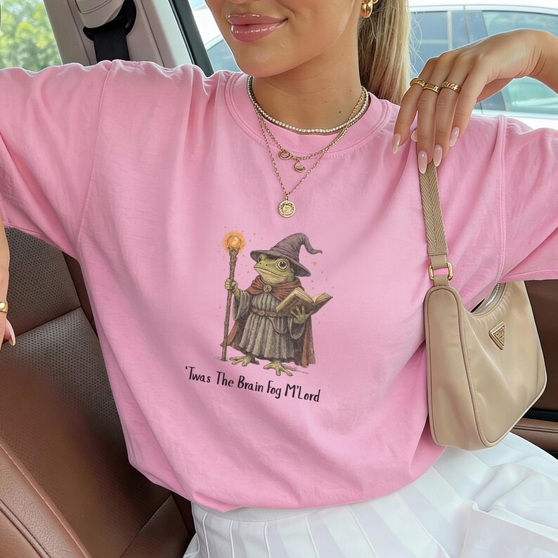 K&ouml;nnte beinhalten: Hellrosa T-Shirt mit einer Illustration eines Froschzauberers, der einen Stab und ein Buch h&auml;lt. Der Frosch tr&auml;gt einen Zaubererhut und einen Umhang. Der Text "'Twas The Brain Fog M'Lord" ist darunter gedruckt.