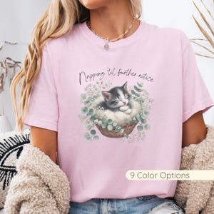 May include: Pink t-shirt featuring a watercolor illustration of a sleeping gray and white kitten in a woven basket surrounded by greenery. The text "Napping 'til further notice" is above the kitten. The shirt also has "9 Color Options" text.