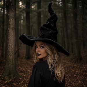 May include: A black velvet witch hat with a tall, pointed, and slightly slumped crown and a wide, curved brim. The fabric has a soft, textured appearance that catches the light, creating subtle highlights across the surface of the hat.