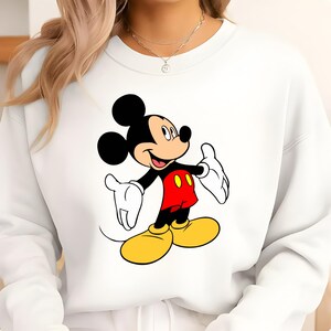 K&ouml;nnte beinhalten: Wei&szlig;es Sweatshirt mit einer Mickey Mouse-Grafik. Die Zeichentrickmaus wird mit schwarzen Ohren, einem schwarzen K&ouml;rper, roten Shorts, gelben Schuhen und wei&szlig;en Handschuhen gezeigt. Das Sweatshirt ist ein l&auml;ssiger Stil.