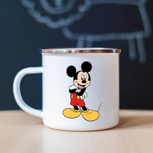 K&ouml;nnte beinhalten: Wei&szlig;er Emaillebecher mit einem Mickey Mouse-Design. Die Zeichentrickmaus ist in ihrer ikonischen Kleidung dargestellt: rote Shorts, gelbe Schuhe und schwarze Handschuhe. Der Becher hat einen silbernen Rand und einen wei&szlig;en Griff.