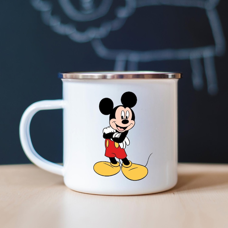 K&ouml;nnte beinhalten: Wei&szlig;er Emaillebecher mit einem Mickey Mouse-Design. Die Zeichentrickmaus ist in ihrer ikonischen Kleidung dargestellt: rote Shorts, gelbe Schuhe und schwarze Handschuhe. Der Becher hat einen silbernen Rand und einen wei&szlig;en Griff.