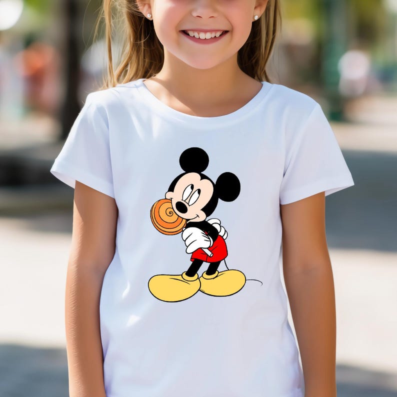 K&ouml;nnte beinhalten: Wei&szlig;es T-Shirt mit einem Cartoon-Motiv von Mickey Mouse, der einen Lolli h&auml;lt. Mickey Mouse ist in seiner klassischen Kleidung dargestellt: schwarze Ohren, rote Shorts, gelbe Schuhe und wei&szlig;e Handschuhe. Das Shirt hat kurze &Auml;rmel und scheint aus Baumwolle zu sein.