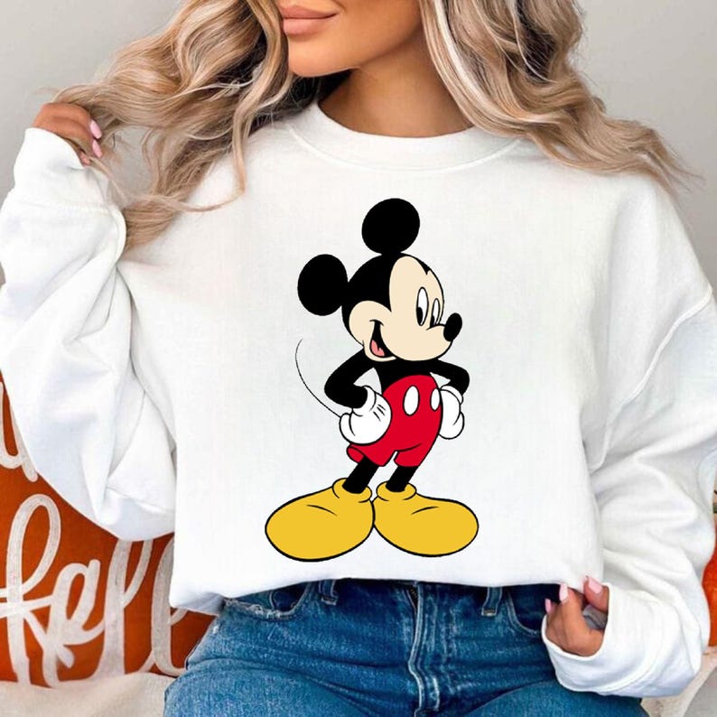 K&ouml;nnte beinhalten: Wei&szlig;es Sweatshirt mit einem farbenfrohen Mickey Mouse-Motiv. Mickey ist in seiner klassischen Kleidung dargestellt: rote Shorts, gelbe Schuhe, schwarze Handschuhe und ein schwarzer K&ouml;rper. Das Sweatshirt ist ein l&auml;ssiger, bequemer Stil.