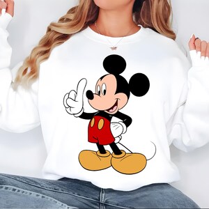 K&ouml;nnte beinhalten: Wei&szlig;es Sweatshirt mit einer Cartoon-Illustration von Mickey Mouse. Mickey wird mit schwarzen Ohren, roten Shorts, gelben Schuhen und wei&szlig;en Handschuhen dargestellt, die nach oben zeigen. Das Sweatshirt ist ein l&auml;ssiger Stil.