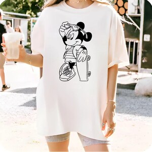 May include: Cream-colored t-shirt featuring a black and white graphic of Mickey Mouse in sunglasses, holding a skateboard. Mickey is wearing a striped shirt and sneakers. The shirt is a relaxed fit.