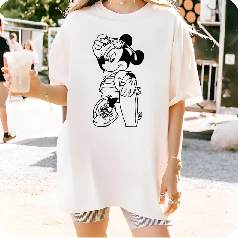 May include: Cream-colored t-shirt featuring a black and white graphic of Mickey Mouse in sunglasses, holding a skateboard. Mickey is wearing a striped shirt and sneakers. The shirt is a relaxed fit.
