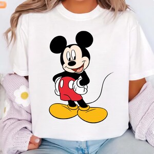 K&ouml;nnte beinhalten: Wei&szlig;es T-Shirt mit einem Cartoon-Bild von Mickey Mouse. Mickey Mouse hat schwarze Ohren, einen schwarzen K&ouml;rper, rote Shorts, gelbe Schuhe und wei&szlig;e Handschuhe. Die Cartoon-Figur l&auml;chelt und streckt die Zunge heraus.