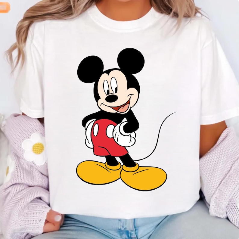 K&ouml;nnte beinhalten: Wei&szlig;es T-Shirt mit einem Cartoon-Bild von Mickey Mouse. Mickey Mouse hat schwarze Ohren, einen schwarzen K&ouml;rper, rote Shorts, gelbe Schuhe und wei&szlig;e Handschuhe. Die Cartoon-Figur l&auml;chelt und streckt die Zunge heraus.