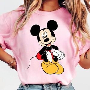 K&ouml;nnte beinhalten: Ein hellrosa T-Shirt mit einer l&auml;chelnden Mickey Mouse-Grafik. Mickey tr&auml;gt seine klassische Kleidung: schwarze Shorts, rote Shorts, gelbe Schuhe und wei&szlig;e Handschuhe. Die Zeichentrickfigur sitzt mit den H&auml;nden auf dem Scho&szlig;.