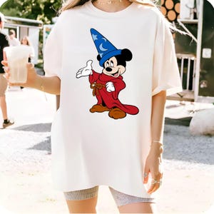 K&ouml;nnte beinhalten: Cremefarbenes T-Shirt mit einer Cartoon-Illustration von Mickey Mouse als Zauberer. Mickey tr&auml;gt einen blauen Hut mit Mond und Sternen, einen roten Umhang und braune Schuhe. Das Design ist mittig auf dem Shirt platziert.
