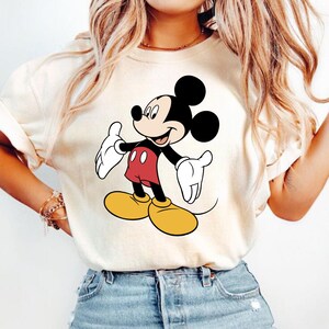 K&ouml;nnte beinhalten: Cremefarbenes T-Shirt mit einer l&auml;chelnden Mickey Mouse-Grafik. Mickey ist in seiner klassischen Kleidung dargestellt: rote Shorts, gelbe Schuhe, wei&szlig;e Handschuhe und schwarze Ohren. Das Shirt ist ein l&auml;ssiger Stil, geeignet f&uuml;r den Alltag.