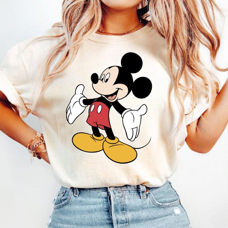 K&ouml;nnte beinhalten: Cremefarbenes T-Shirt mit einer l&auml;chelnden Mickey Mouse-Grafik. Mickey ist in seiner klassischen Kleidung dargestellt: rote Shorts, gelbe Schuhe, wei&szlig;e Handschuhe und schwarze Ohren. Das Shirt ist ein l&auml;ssiger Stil, geeignet f&uuml;r den Alltag.