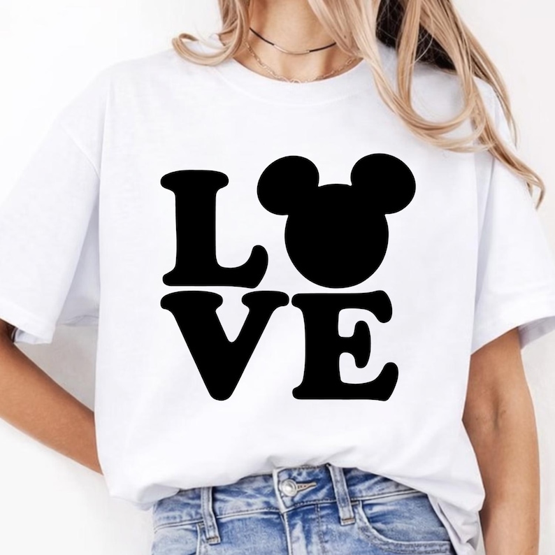 Puede incluir: Camiseta blanca con la palabra "LOVE" en letras negras y una cabeza de Mickey Mouse negra que reemplaza la "O". La camiseta es de estilo casual.