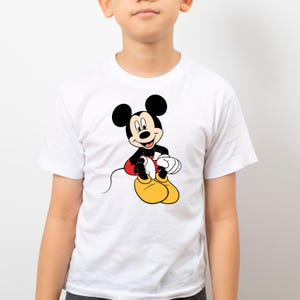 K&ouml;nnte beinhalten: Wei&szlig;es T-Shirt mit einem Mickey Mouse-Cartoon-Motiv. Mickey ist in seiner klassischen Kleidung dargestellt: schwarze Ohren, rote Shorts, gelbe Schuhe und wei&szlig;e Handschuhe. Das T-Shirt hat kurze &Auml;rmel und ist wahrscheinlich aus Baumwolle.