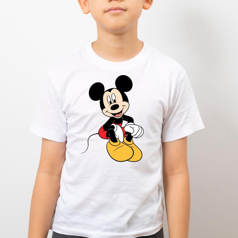 K&ouml;nnte beinhalten: Wei&szlig;es T-Shirt mit einem Mickey Mouse-Cartoon-Motiv. Mickey ist in seiner klassischen Kleidung dargestellt: schwarze Ohren, rote Shorts, gelbe Schuhe und wei&szlig;e Handschuhe. Das T-Shirt hat kurze &Auml;rmel und ist wahrscheinlich aus Baumwolle.