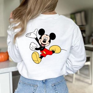K&ouml;nnte beinhalten: Wei&szlig;es Sweatshirt mit einem farbenfrohen Mickey Mouse-Motiv auf dem R&uuml;cken. Die Cartoonfigur ist in Bewegung dargestellt, mit roter Hose, gelben Schuhen und schwarzen Handschuhen. Das Sweatshirt hat lange &Auml;rmel.