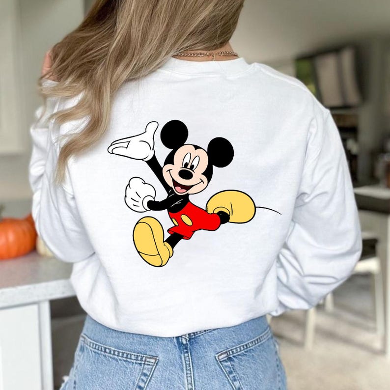 K&ouml;nnte beinhalten: Wei&szlig;es Sweatshirt mit einem farbenfrohen Mickey Mouse-Motiv auf dem R&uuml;cken. Die Cartoonfigur ist in Bewegung dargestellt, mit roter Hose, gelben Schuhen und schwarzen Handschuhen. Das Sweatshirt hat lange &Auml;rmel.