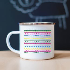 May include: White enamel mug with a silver rim and handle. The mug features a colourful geometric pattern in green, pink, yellow, and blue. The design includes lines, zigzags, and other shapes.