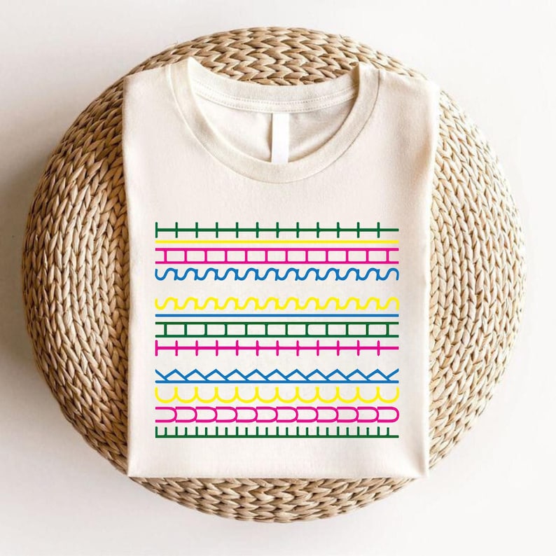 May include: A cream-coloured t-shirt with a vibrant geometric pattern. The design incorporates horizontal lines in green, pink, yellow, and blue, with diverse shapes such as zigzags and curves. The shirt is folded on a woven, circular surface.