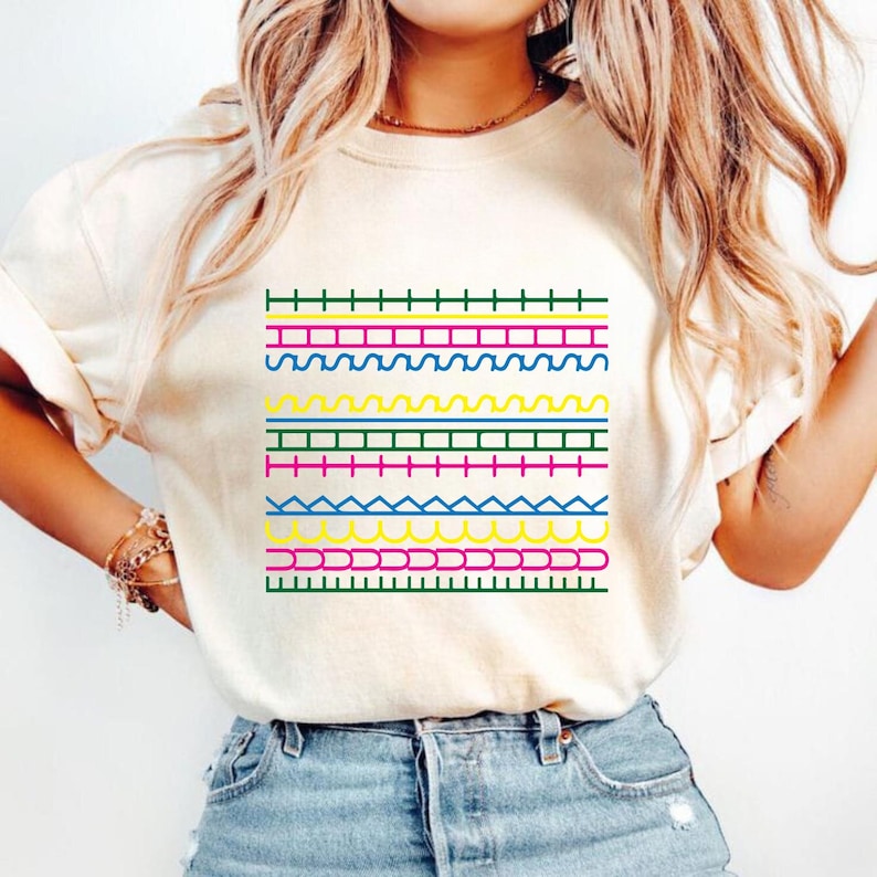 May include: Cream-coloured t-shirt with a vibrant geometric design. The design features horizontal lines in green, blue, yellow, pink, and red. The shirt is made of a soft material, perfect for casual wear. The design is eye-catching.