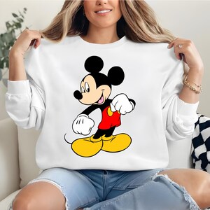 K&ouml;nnte beinhalten: Wei&szlig;es Sweatshirt mit einer Mickey Mouse-Grafik. Die Zeichentrickfigur ist in ihrer ikonischen Kleidung dargestellt: rote Shorts, gelbe Schuhe, wei&szlig;e Handschuhe und schwarze Ohren. Das Sweatshirt ist ein l&auml;ssiger, bequemer Stil.