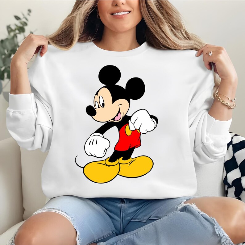 K&ouml;nnte beinhalten: Wei&szlig;es Sweatshirt mit einer Mickey Mouse-Grafik. Die Zeichentrickfigur ist in ihrer ikonischen Kleidung dargestellt: rote Shorts, gelbe Schuhe, wei&szlig;e Handschuhe und schwarze Ohren. Das Sweatshirt ist ein l&auml;ssiger, bequemer Stil.