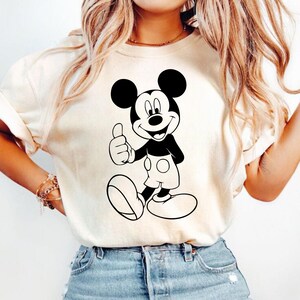 May include: Cream-colored t-shirt featuring a black and white graphic of Mickey Mouse giving a thumbs-up. The cartoon character is smiling and wearing white shorts. The shirt is a casual style.