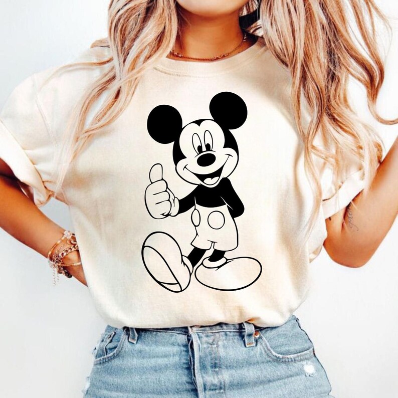 May include: Cream-colored t-shirt featuring a black and white graphic of Mickey Mouse giving a thumbs-up. The cartoon character is smiling and wearing white shorts. The shirt is a casual style.