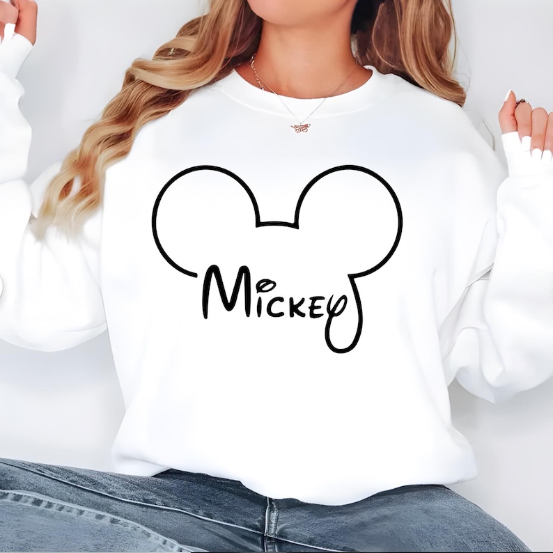 Puede incluir: Sudadera blanca con un contorno negro de las orejas de Mickey Mouse y el nombre "Mickey" en cursiva. La sudadera es de color blanco liso.