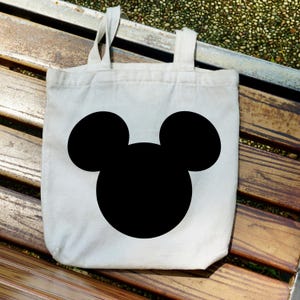 Puede incluir: Un bolso tote de lona blanca con una gran silueta negra de la cabeza de Mickey Mouse. El bolso tiene dos asas blancas y est&aacute; sobre un banco de madera.