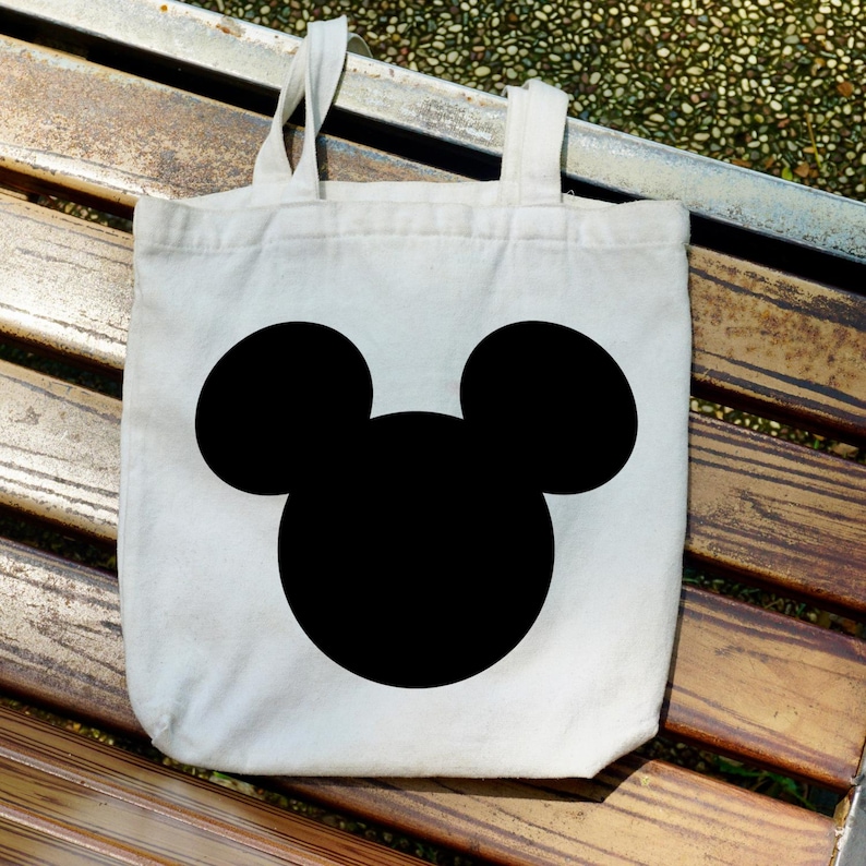 Puede incluir: Un bolso tote de lona blanca con una gran silueta negra de la cabeza de Mickey Mouse. El bolso tiene dos asas blancas y est&aacute; sobre un banco de madera.