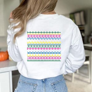 May include: White sweatshirt with a colourful geometric design on the back. The design features horizontal rows of various shapes and colours, including green, pink, yellow, and blue. A casual, everyday garment.