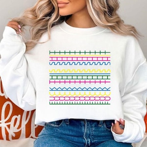 May include: White sweatshirt with a vibrant geometric pattern. The design incorporates horizontal rows of green, pink, yellow, blue, and pink lines in diverse patterns. A casual, comfortable style of clothing.