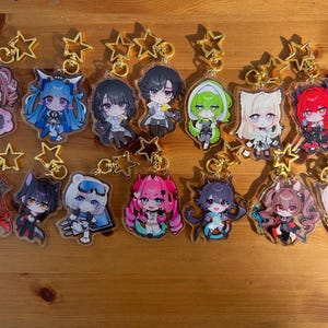 May include: Collection of anime-style character keychains with gold star-shaped clasps. Each keychain features a unique character design with vibrant colors and detailed artwork. The keychains are arranged on a wooden surface.
